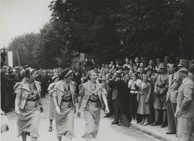 Vier deelneemsters passeren te Wijchen op de tweede dag van de 27e Vierdaagse ZKH Prins Bernhard (met anjer). Hij wordt geflankeerd door de kapitein E.C. baron van Heeckeren van Brandsenburg en (half zichtbaar) de kapitein J.N. Breunese. Achter de fotograaf, de journalist H.A. Meerum Terwogt (met gleufhoed en insigne).